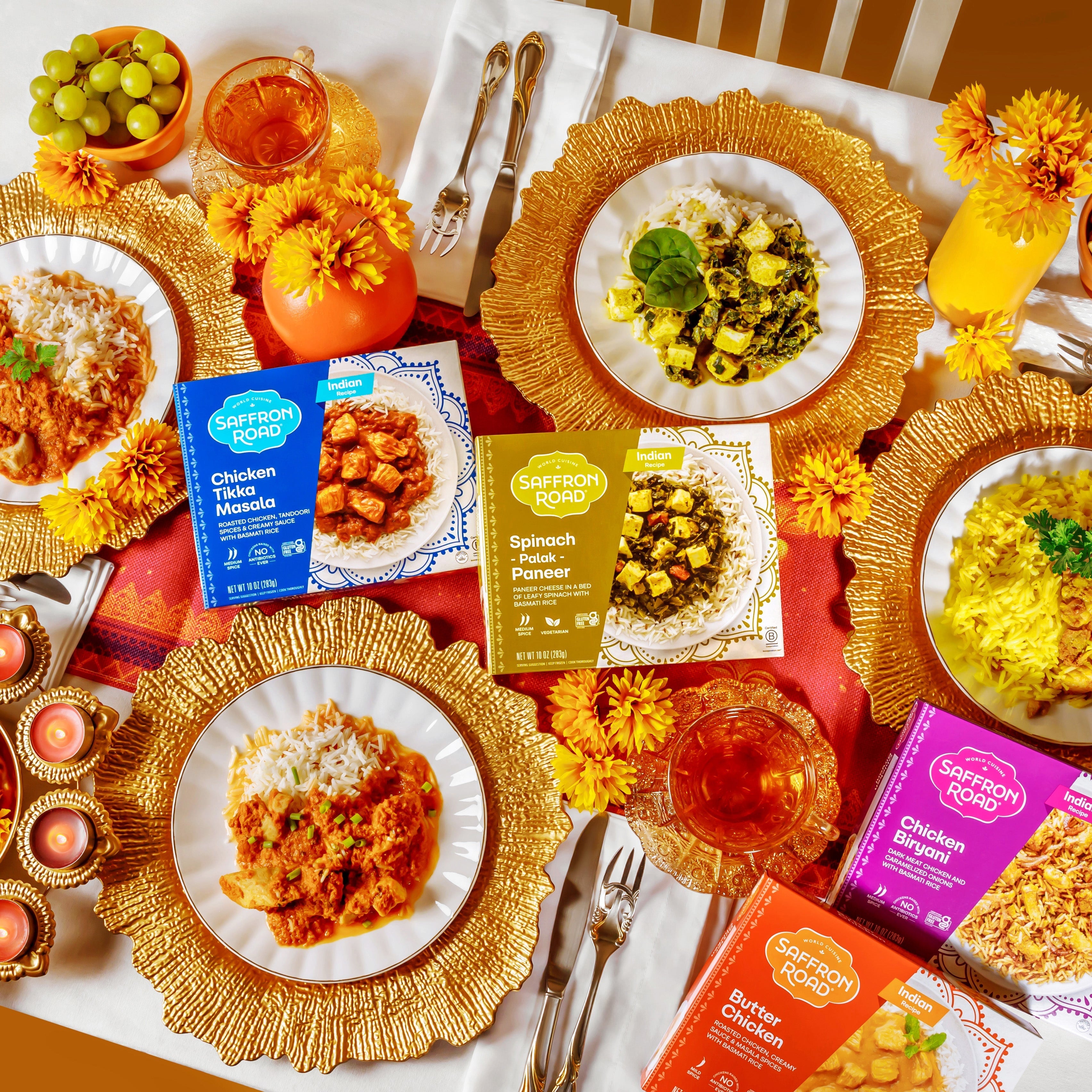 Butter Chicken Frozen Meal package, Chicken Biryani package, Chicken Tikka Masala package, Spinach Palak Paneer package all with corresponding meals prepared and plated on a table with Diwali decorations of gold, orange, and red