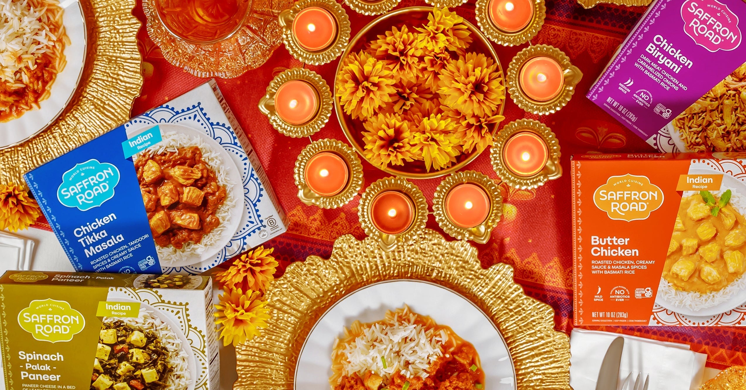 A festive table setting featuring Saffron Road frozen Indian entrees including Chicken Tikka Masala, Butter Chicken, Chicken Biryani, and Spinach Palak Paneer. The table is adorned with golden plates, marigold flowers, and traditional candles, evoking a celebration atmosphere.